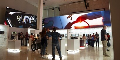 Trade show floor with illuminated product kiosks, attendees chatting in small groups, and a large digital display showing EV charging equipment overhead.
